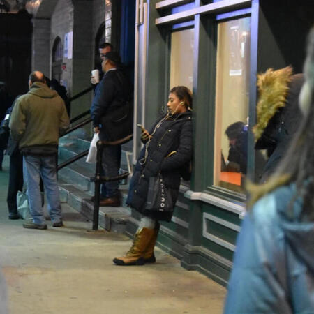 hoboken_mlleone A woman in a long dark coat leans against a wall in the street, on her phone.