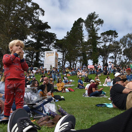 hsbcrowd_mlleone A crowd of people sit on a grassy hill, including a little child wearing red pajamas.