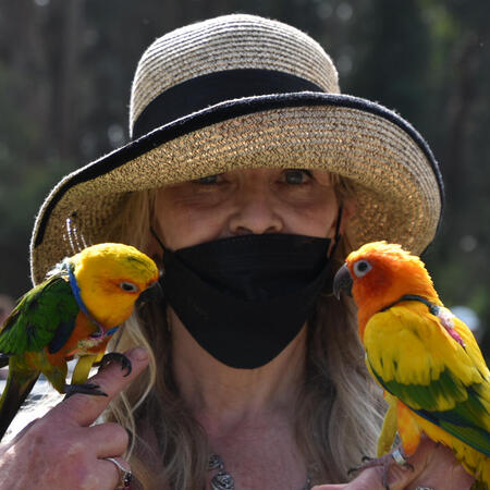 hsbparrots_mlleone A woman in a straw hat holds two yellow and green parrots and looks directly into the camera.