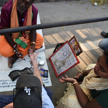 indiapalm_mlleone A man in sits and reads the palm of another man in a baseball cap while a woman translate for him.