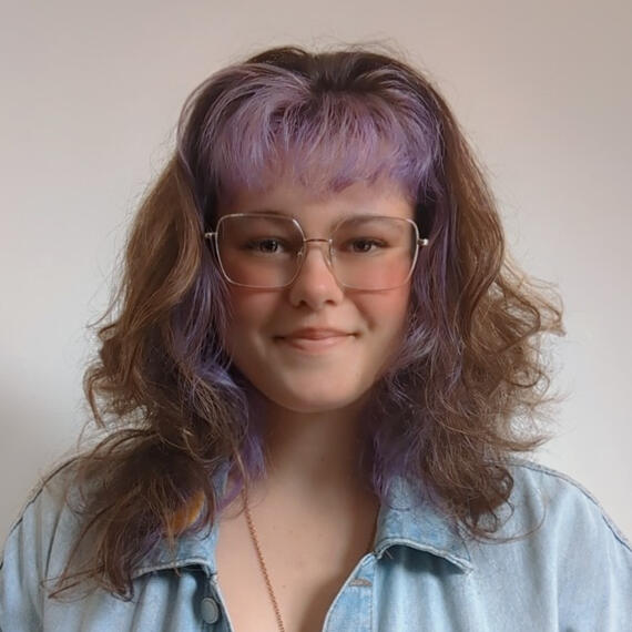 A headshot of Marie Louise Leone, a white woman with brown curly hair dyed purple in the front and wire-rimmed glasses.