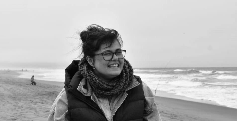 coverphoto_mlleone A black and white photo of Marie Louise "Marlo" Leone, a white woman with dark hair and glasses. She's standing on a beach, wearing a jacket, vest and scarf and smiling.